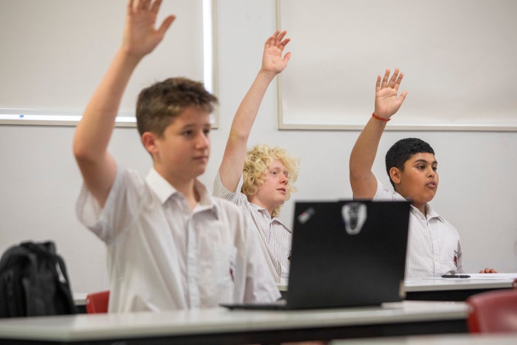 3 students engaging in class learning and putting their hands up to answer a question.