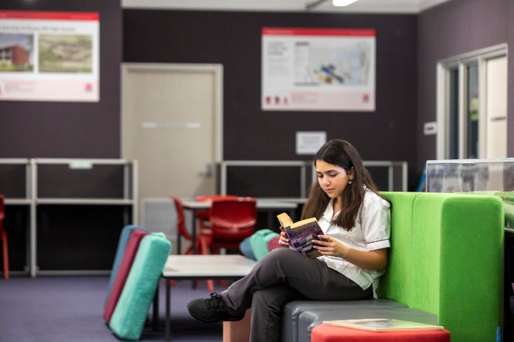 A student reading a book on a green lounge within a school library.