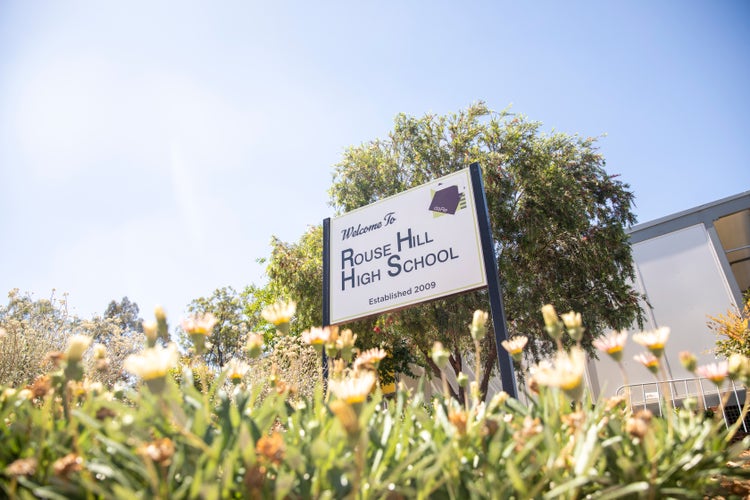 A 'Welcome to Rouse Hill High School' sign infront of a large green tree with flowers in the foreground.