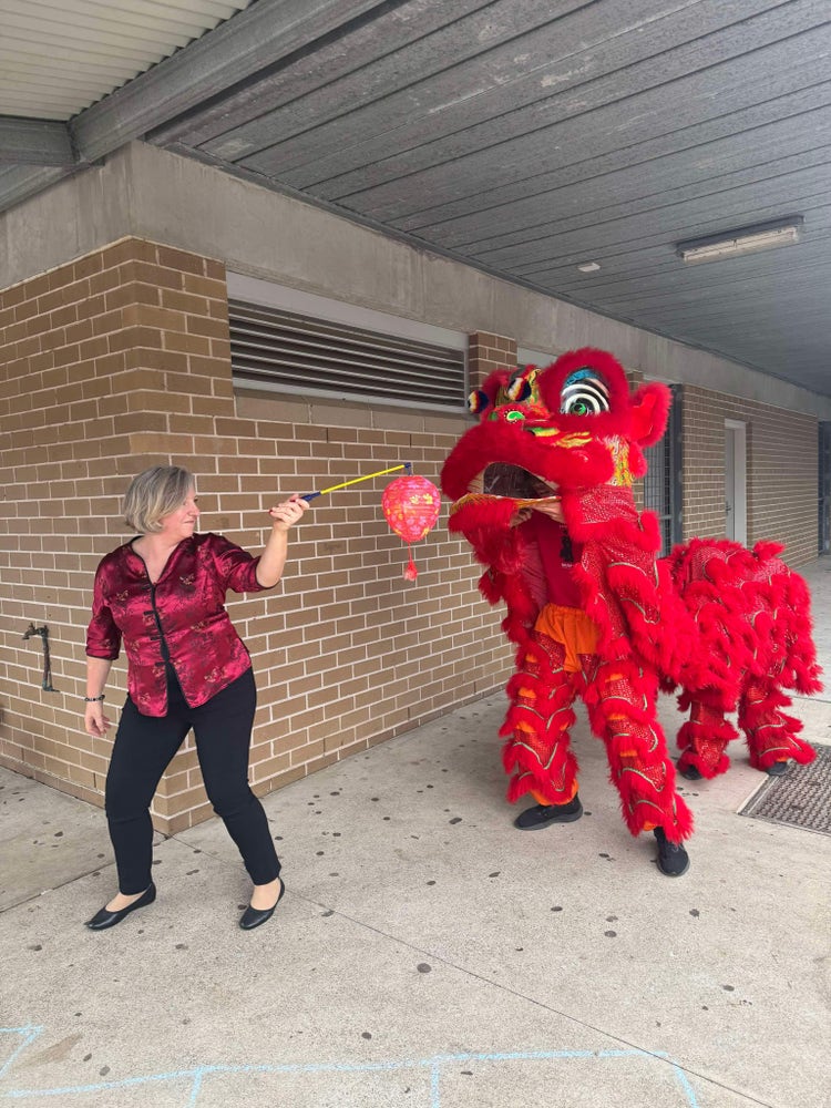 A teacher wearing a red shirt and black pants holding a red lantern while a red chinese dragon costume character is following her.