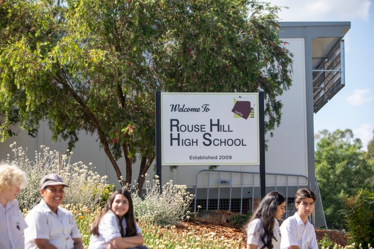 A group of students sitting infront of the school entrance sign